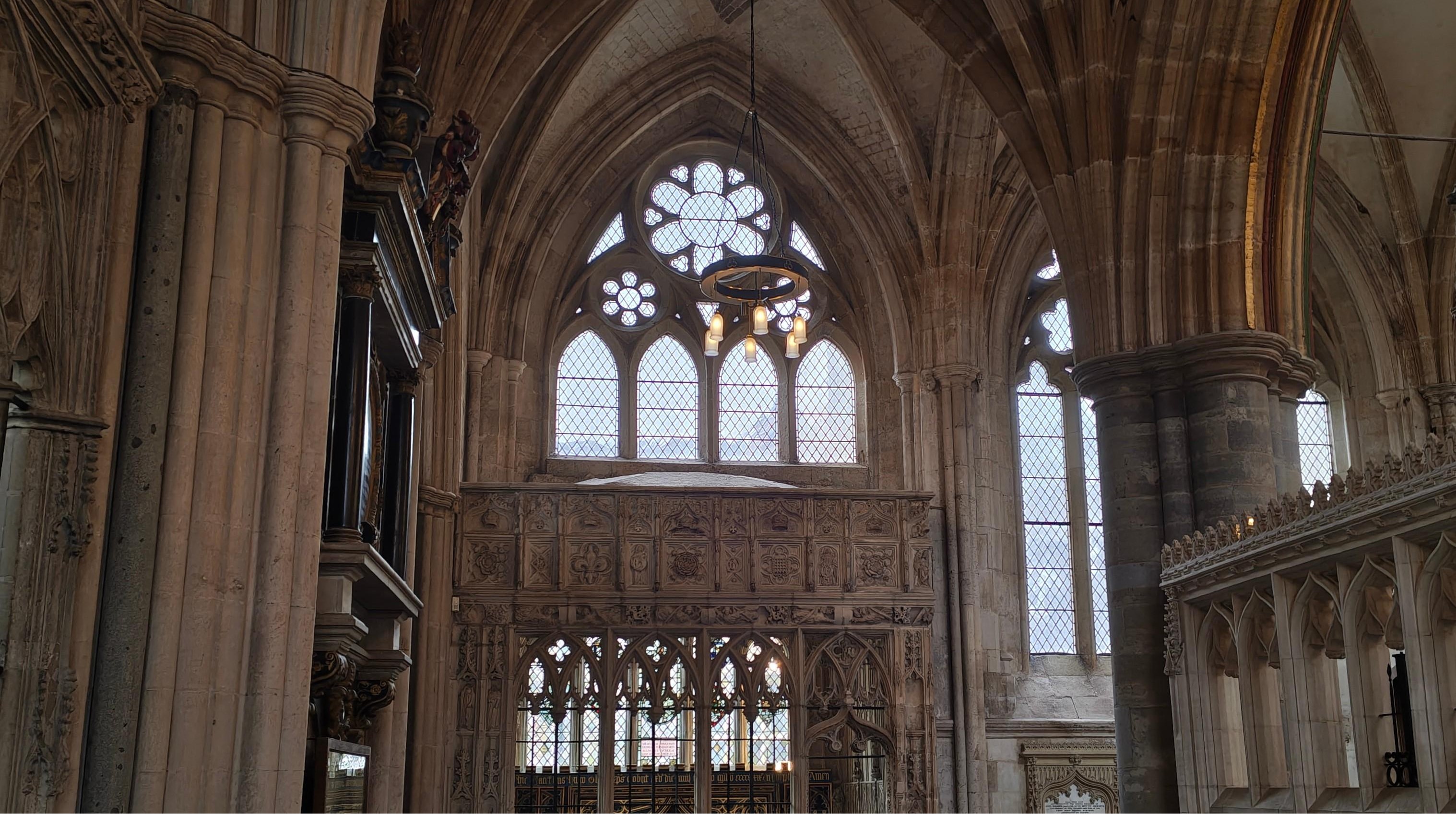 Exeter-Cathedral-Window-3035×1699 The intricate interior of Exeter Cathedral, featuring a Gothic stone window with circular rose tracery, vaulted arches, and an ornate carved stone screen bathed in soft natural light.
