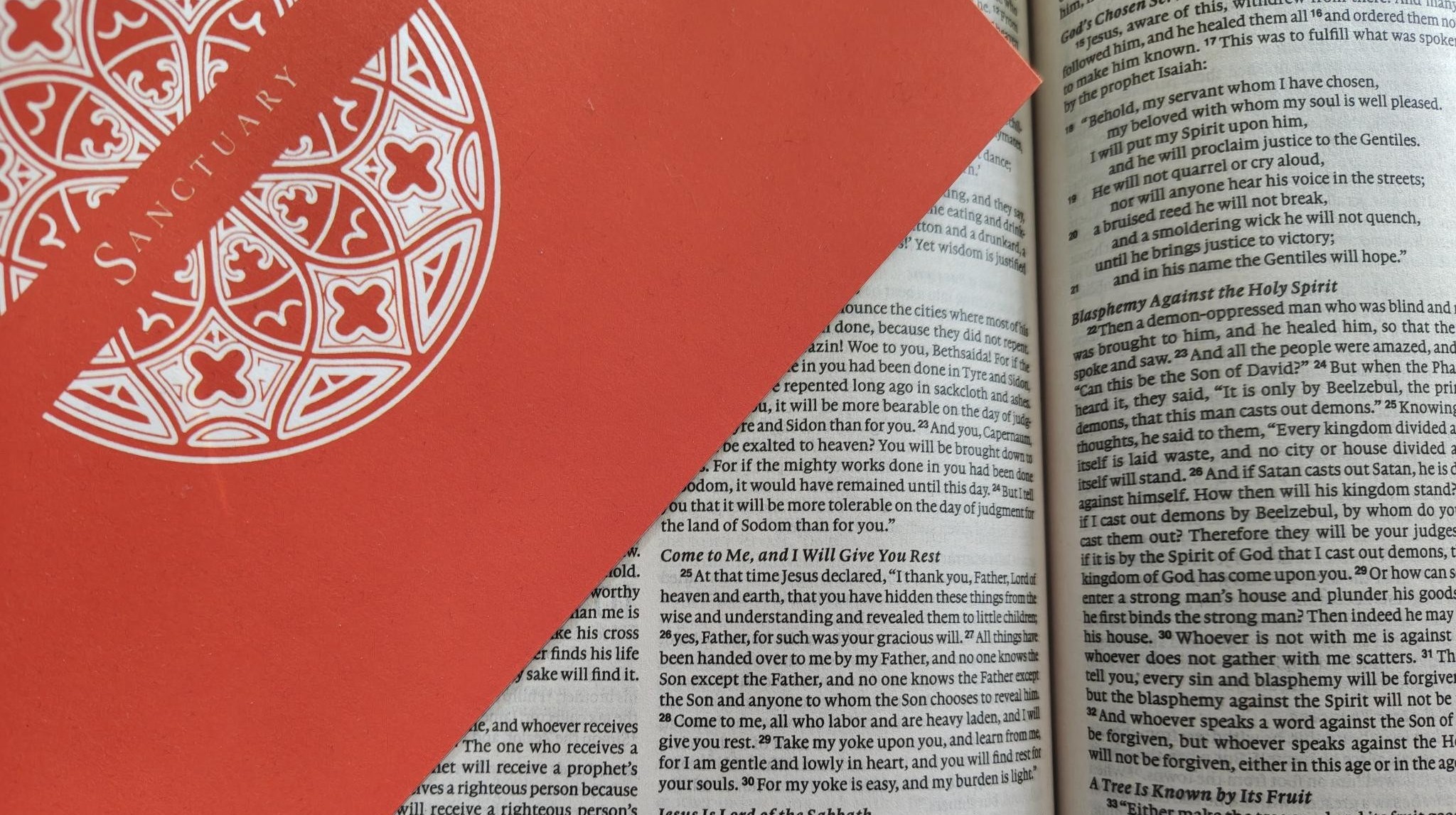 Matthew-11-28-Sanctuary-Card-Bible-Quote A close-up photograph of a vibrant orange 'Sanctuary' greeting card featuring a white Gothic rose window design. The card is resting on an open Bible, positioned specifically to highlight the text of Matthew 11:28.