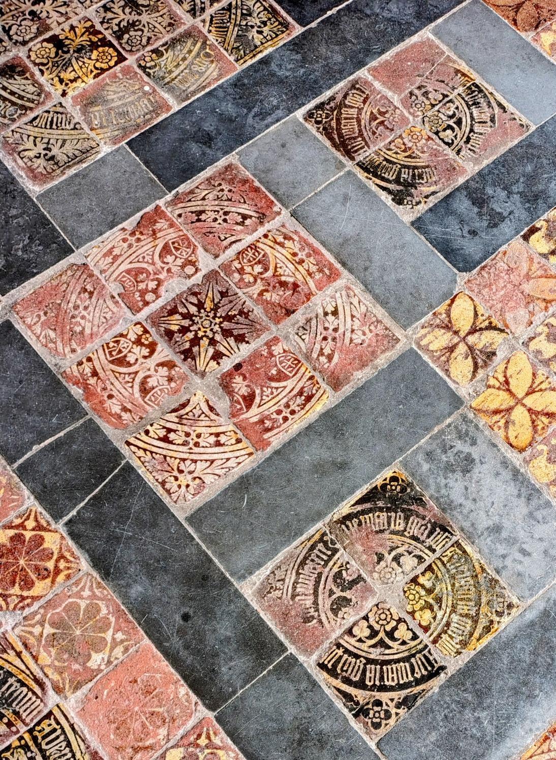 Detailed view of medieval encaustic floor tiles at Gloucester Cathedral, featuring worn geometric and floral patterns in terracotta, gold, and charcoal tones.